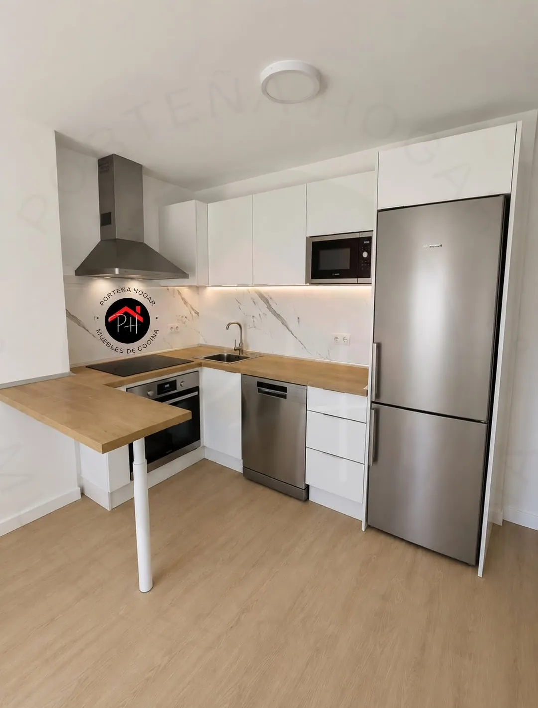 Cocina luminosa en L con muebles blancos, encimera de madera que se prolonga en una barra con pata, frente de aspecto mármol y electrodomésticos de acero